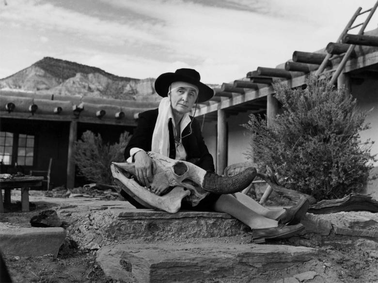 A black-and-white photo of Georgia O'Keeffe sitting in a courtyard before a mountain holding a deer skull.